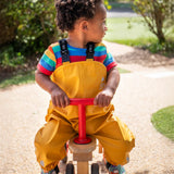 Child riding a bike wearing the Frugi children's waterproof Puddle buster trousers with elasticated arm straps, in bright Bumble Bee Yellow with a rainbow striped t-shirt underneath.