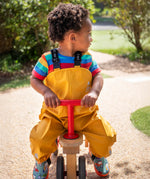 Child riding a bike wearing the Frugi children's waterproof Puddle buster trousers with elasticated arm straps, in bright Bumble Bee Yellow with a rainbow striped t-shirt underneath.