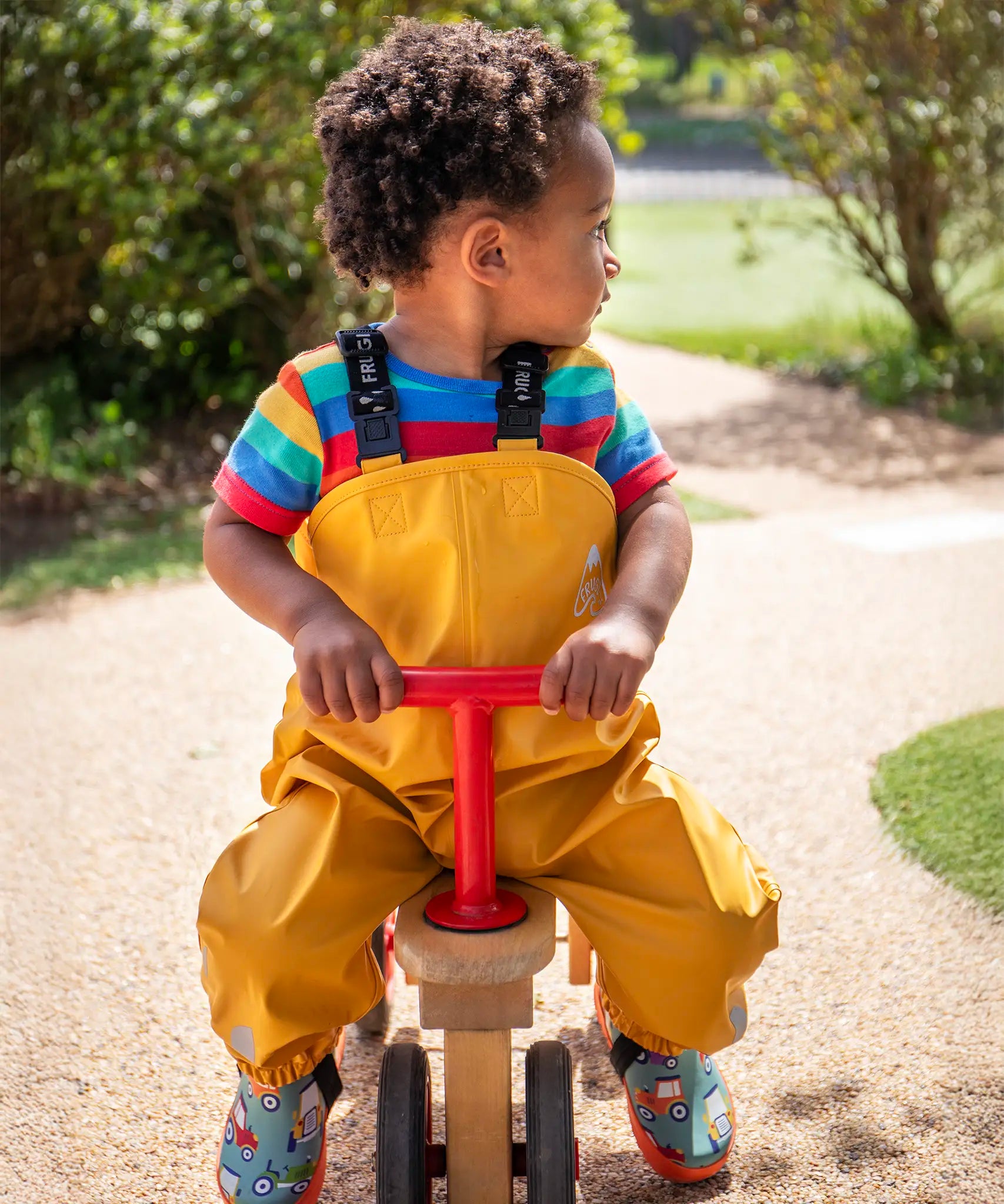 Child riding a bike wearing the Frugi children's waterproof Puddle buster trousers with elasticated arm straps, in bright Bumble Bee Yellow with a rainbow striped t-shirt underneath.