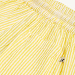 A child happily standing in a green house with their hands in the pockets of the Frugi Archie Seersucker Shorts - Dandelion. A bright yellow and white striped short with an elasticated waist