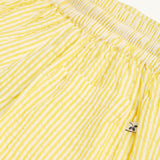 A child happily standing in a green house with their hands in the pockets of the Frugi Archie Seersucker Shorts - Dandelion. A bright yellow and white striped short with an elasticated waist