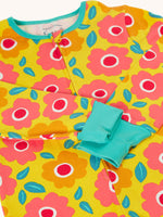 A close up of the front of the Frugi zelah zip up all in one with a floral fields print design. A bright yellow organic cotton all in one with pink and orange flowers all over. The sleeves are shown with the cuffs turned up.  