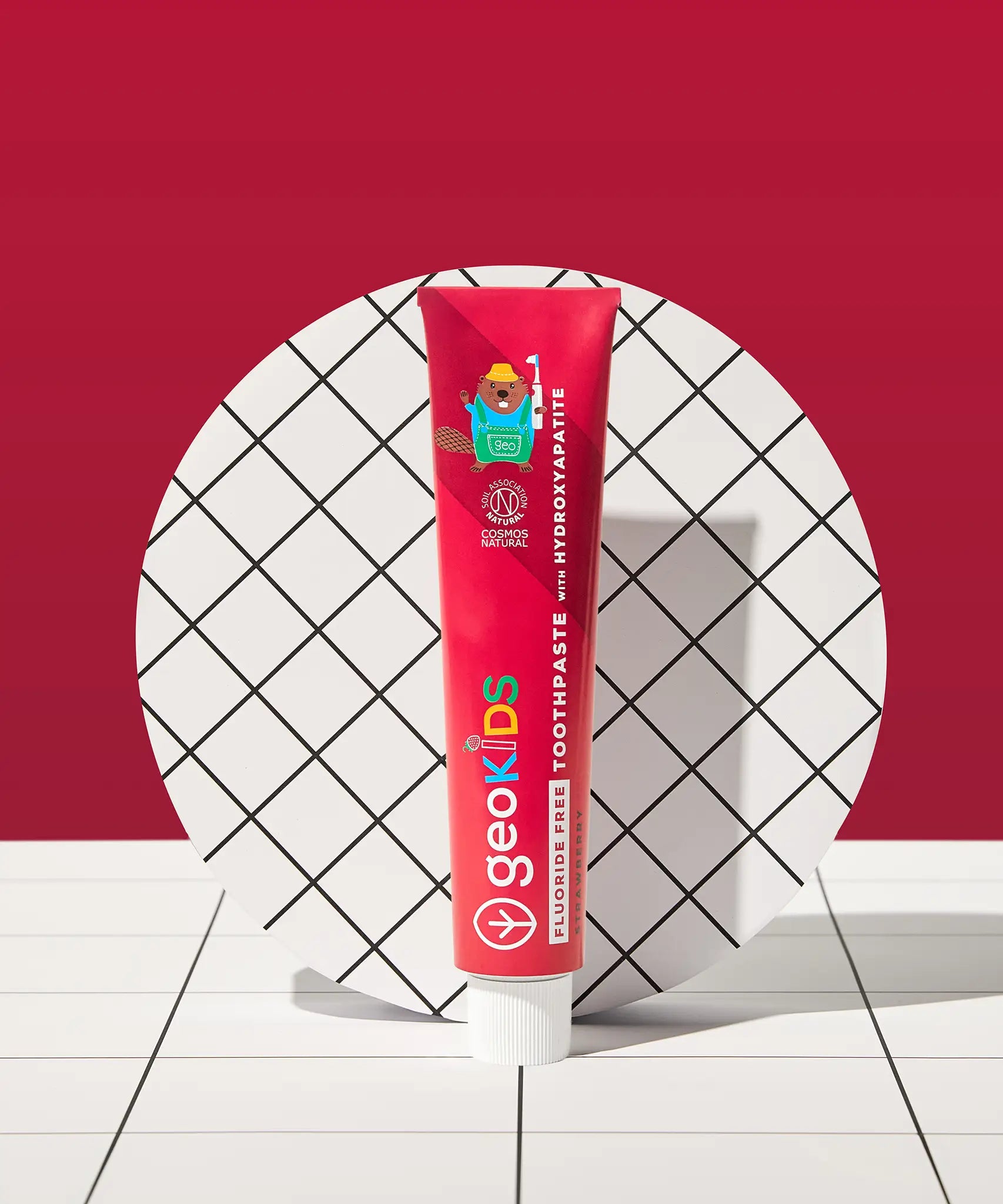 Tube of Georganics strawberry hydroxyapatite toothpaste tube on a tile style floor and matching red background.