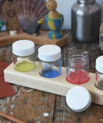 50ml Glass paint jar pots with some  paint inside placed into the circular slots on the wooden holder block. 