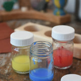 3 Glass paint jar pots with lids with some blue, red and yellow paint in them.