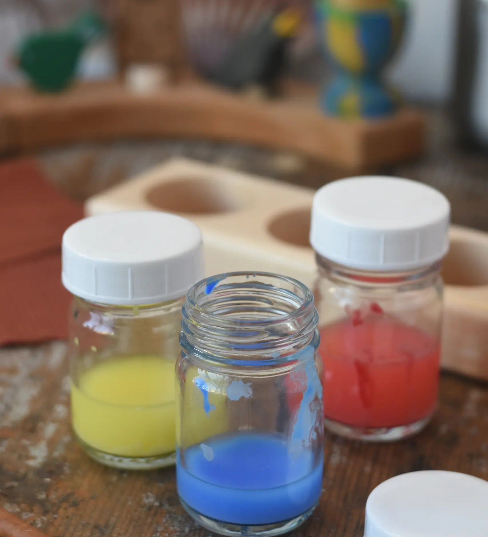 3 Glass paint jar pots with lids with some blue, red and yellow paint in them.