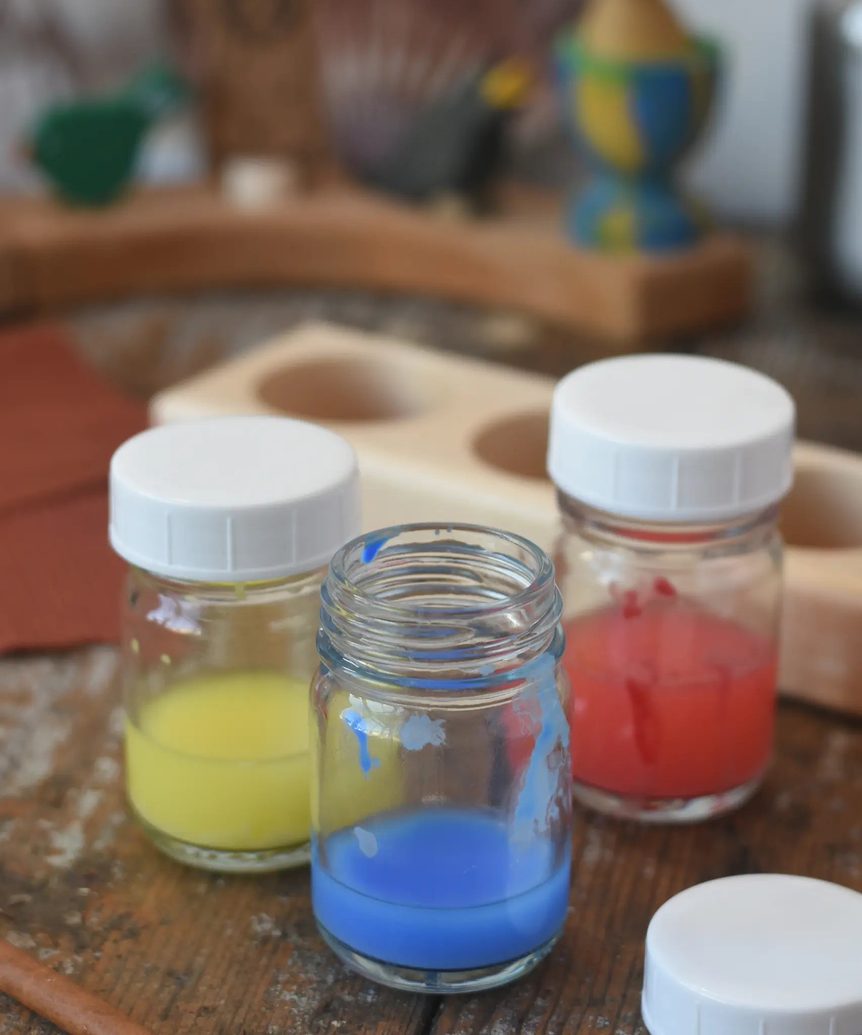 3 Glass paint jar pots with lids with some blue, red and yellow paint in them.