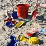 A collection of Gluckskafer sand moulds placed on the sand at a beach. A child's hand can be seen to one side holding a red square shovel.