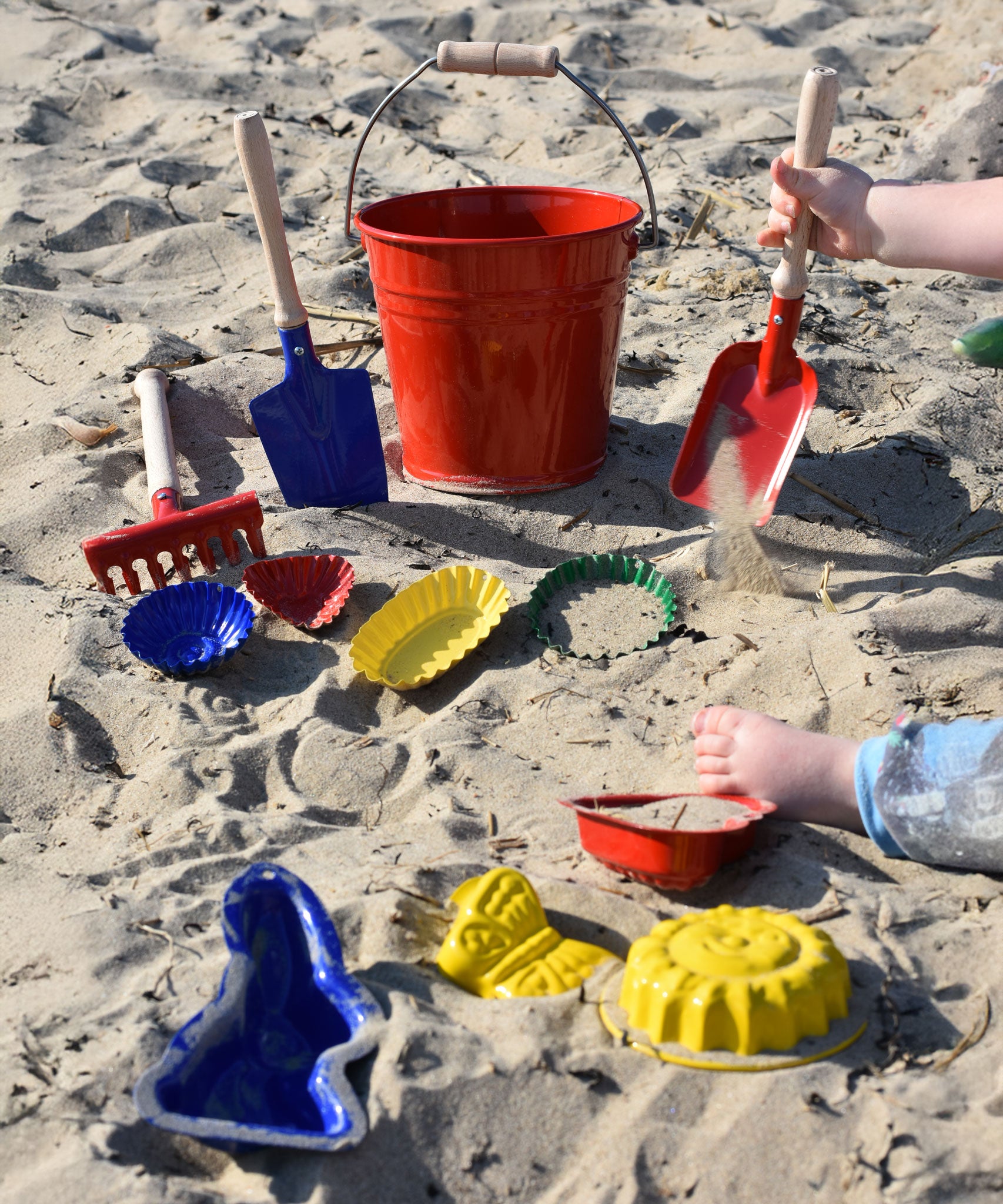 A collection of Gluckskafer sand moulds placed on the sand at a beach. A child's hand can be seen to one side holding a red square shovel.