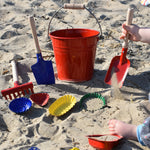 A collection of Glückskäfer Metal outdoor toys and children's tools. A child can be seen to the side using a small red shovel in the sand.  