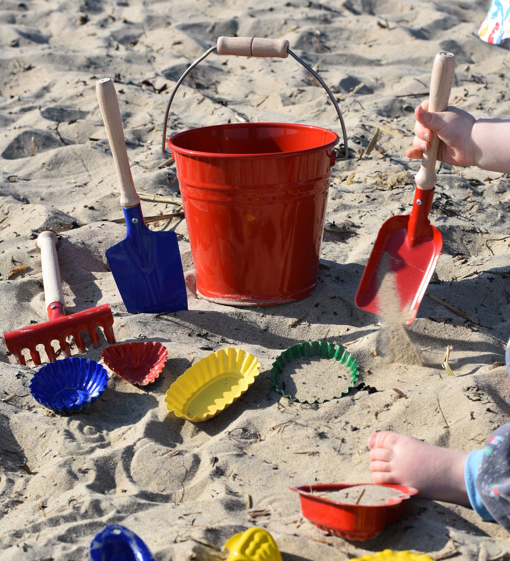 A collection of Glückskäfer Metal outdoor toys and children's tools. A child can be seen to the side using a small red shovel in the sand.  