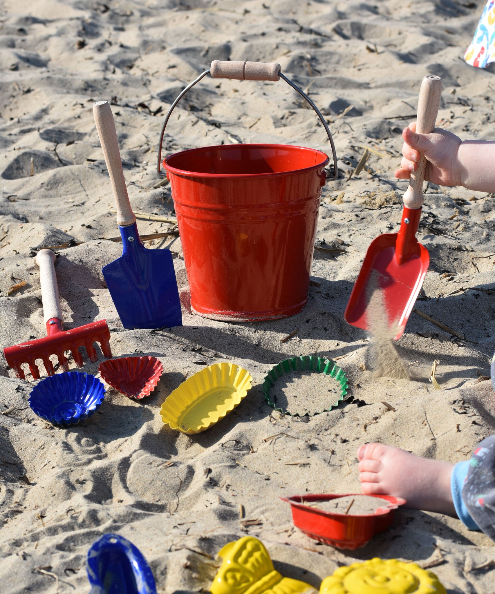 A collection of Glückskäfer Metal outdoor toys and children's tools. A child can be seen to the side using a small red shovel in the sand.  