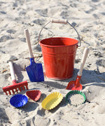 A Glückskäfer Red Metal Bucket placed on the sand at a beach surrounded by metal sand moulds and tools.