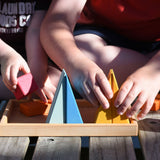 A close up of two children sitting on wooden decking outdoors playing with the Glückskäfer 20-Piece Small Toscana Blocks Set. They are placing various wooden blocs from the set onto the wooden storage display base tray that the set comes with.