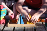 A close up of two children sitting on wooden decking outdoors playing with the Glückskäfer 20-Piece Small Toscana Blocks Set. They are placing various wooden blocs from the set onto the wooden storage display base tray that the set comes with.
