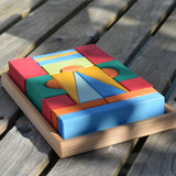 The Glückskäfer 20-Piece Small Toscana Blocks Set placed on wooden decking outdoors. Leafy shadows can be seen covering the wooden building blocks which are perfectly placed n their wooden storage display case. 