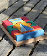 The Glückskäfer 20-Piece Small Toscana Blocks Set placed on wooden decking outdoors. Leafy shadows can be seen covering the wooden building blocks which are perfectly placed n their wooden storage display case. 