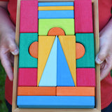 A close up of the Glückskäfer 20-Piece Small Toscana Blocks being held in a child's hands. The set contains various sized wooden building blocks, slates and half cylinder shaped blocks in various colours.