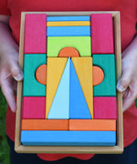 A close up of the Glückskäfer 20-Piece Small Toscana Blocks being held in a child's hands. The set contains various sized wooden building blocks, slates and half cylinder shaped blocks in various colours.