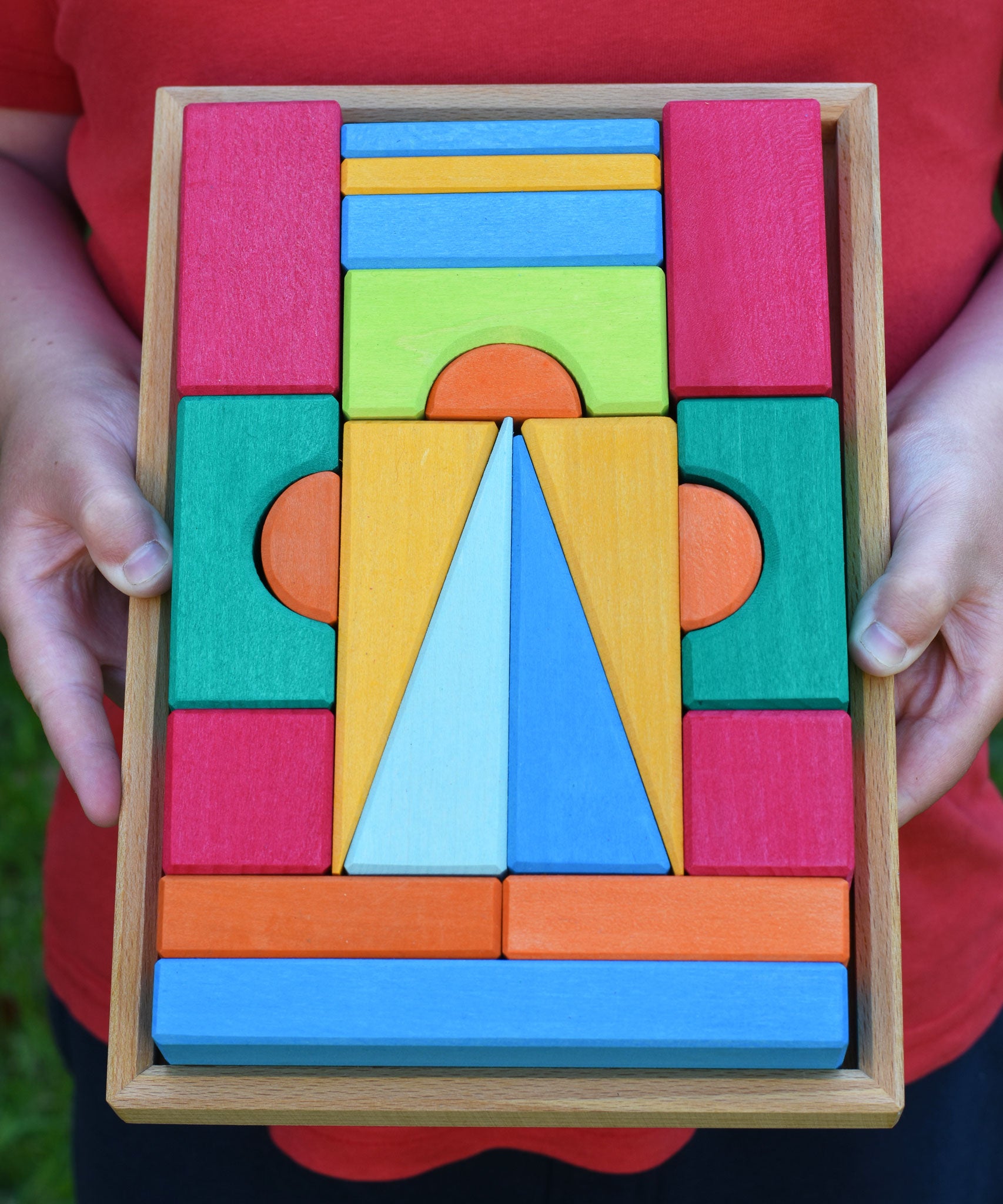 A close up of the Glückskäfer 20-Piece Small Toscana Blocks being held in a child's hands. The set contains various sized wooden building blocks, slates and half cylinder shaped blocks in various colours.