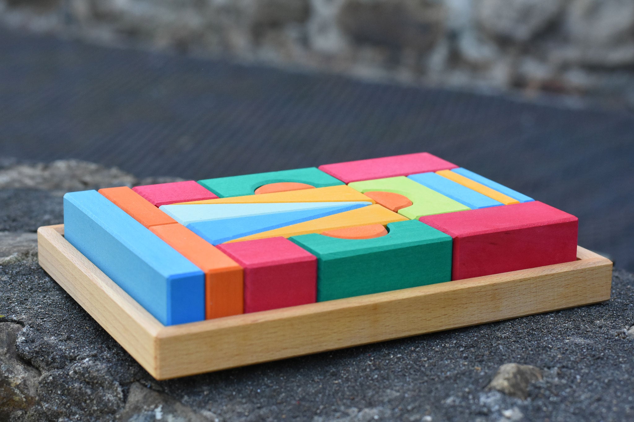 The Glückskäfer 20-Piece Small Toscana Blocks Set shown with the wooden blocks perfectly placed in their wooden storage tray on a stone wall outdoors. 