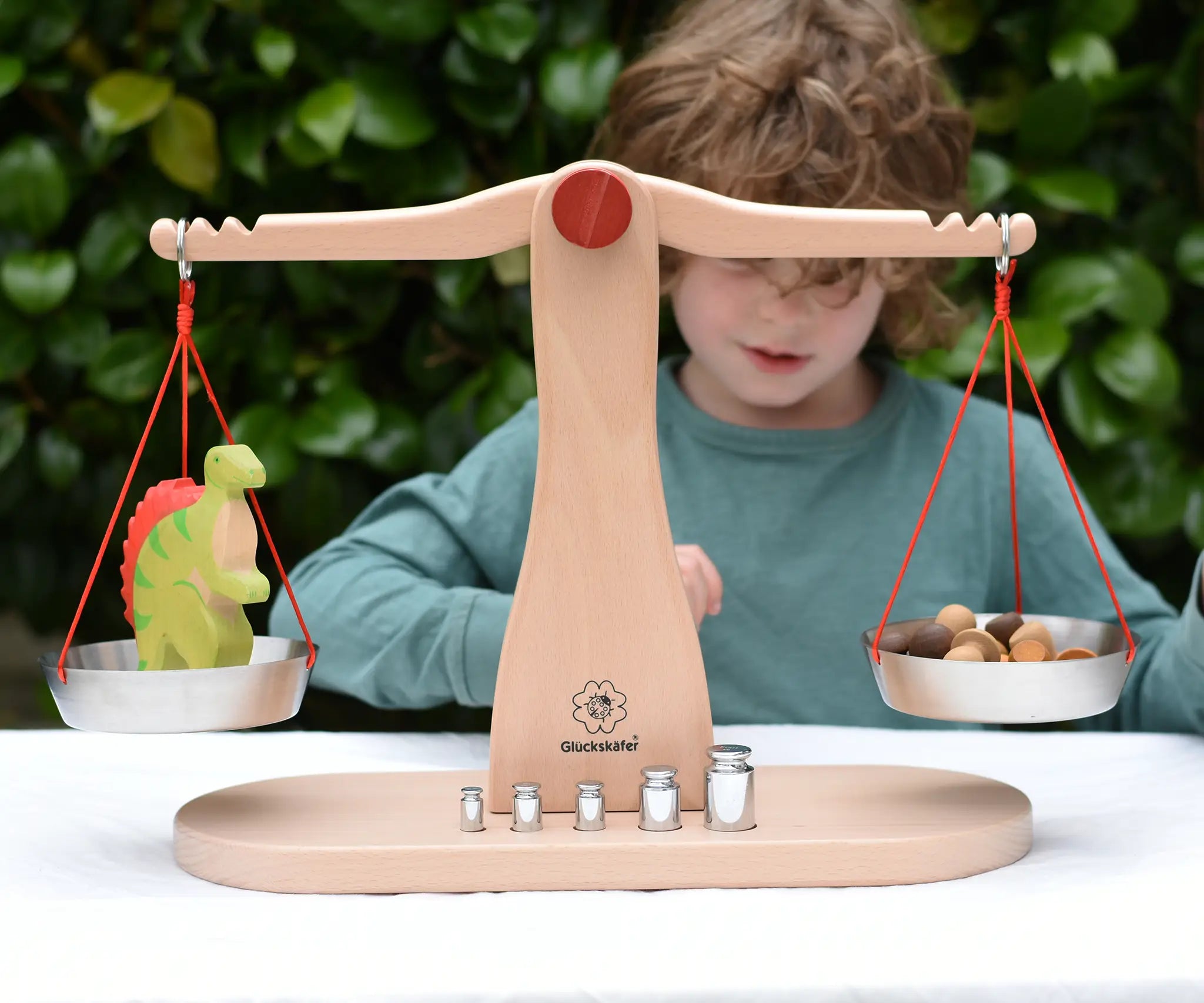 Child playing with the Glückskäfer balance with weights children's weighing scales toy.  One side has a Holztigher spinosaurus dinosaur figure and the other has Grapat mushroom mandala pieces. 