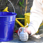 A Glückskäfer Blue Metal Bucket on a slate surface next to a child's hand that is holding onto a seedbom seed pack.