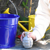 A Glückskäfer Blue Metal Bucket on a slate surface next to a child's hand that is holding onto a seedbom seed pack.