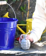 A Glückskäfer Blue Metal Bucket on a slate surface next to a child's hand that is holding onto a seedbom seed pack.
