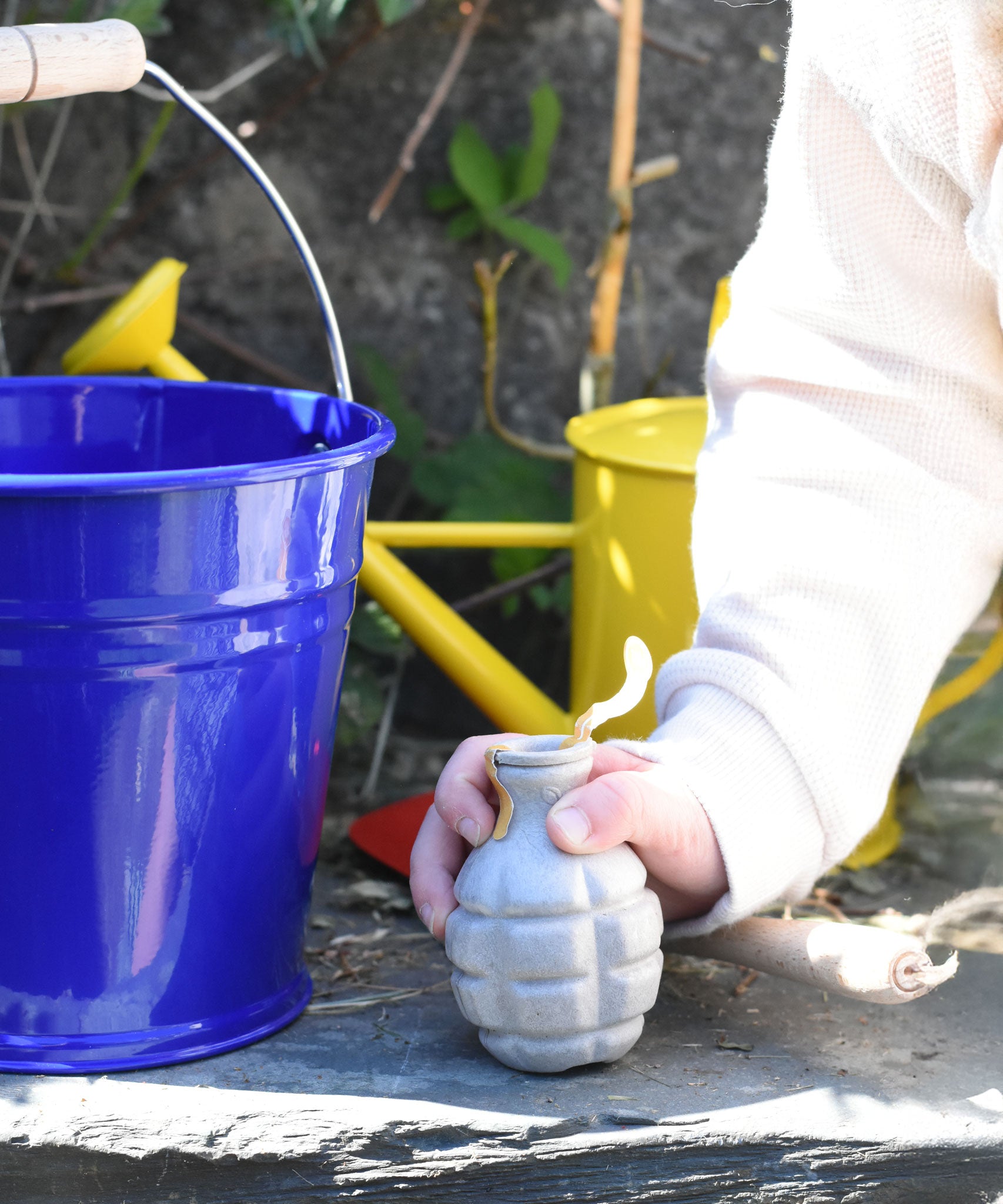 A Glückskäfer Blue Metal Bucket on a slate surface next to a child's hand that is holding onto a seedbom seed pack.