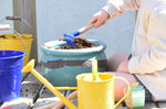 A child using a blue Glückskäfer Metal Blue Rake to prepare the soil in a pot ready for planting.
