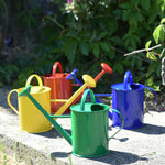 A collection of Glückskäfer Children's 1L Metal Watering Cans. There is a yellow, red, green and blue coloured watering can placed on a concrete surface outdoors. 