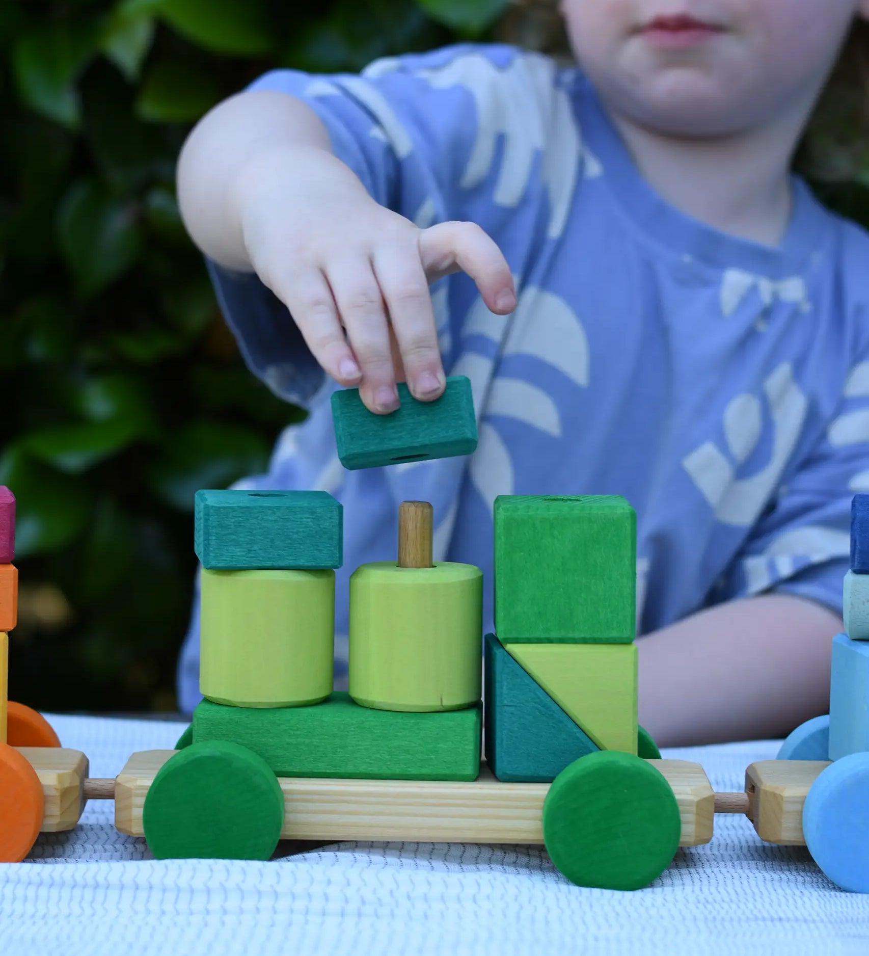 Child playing with the Glückskäfer colourful shape train wooden toy placing a square block on the pillar on the base piece.