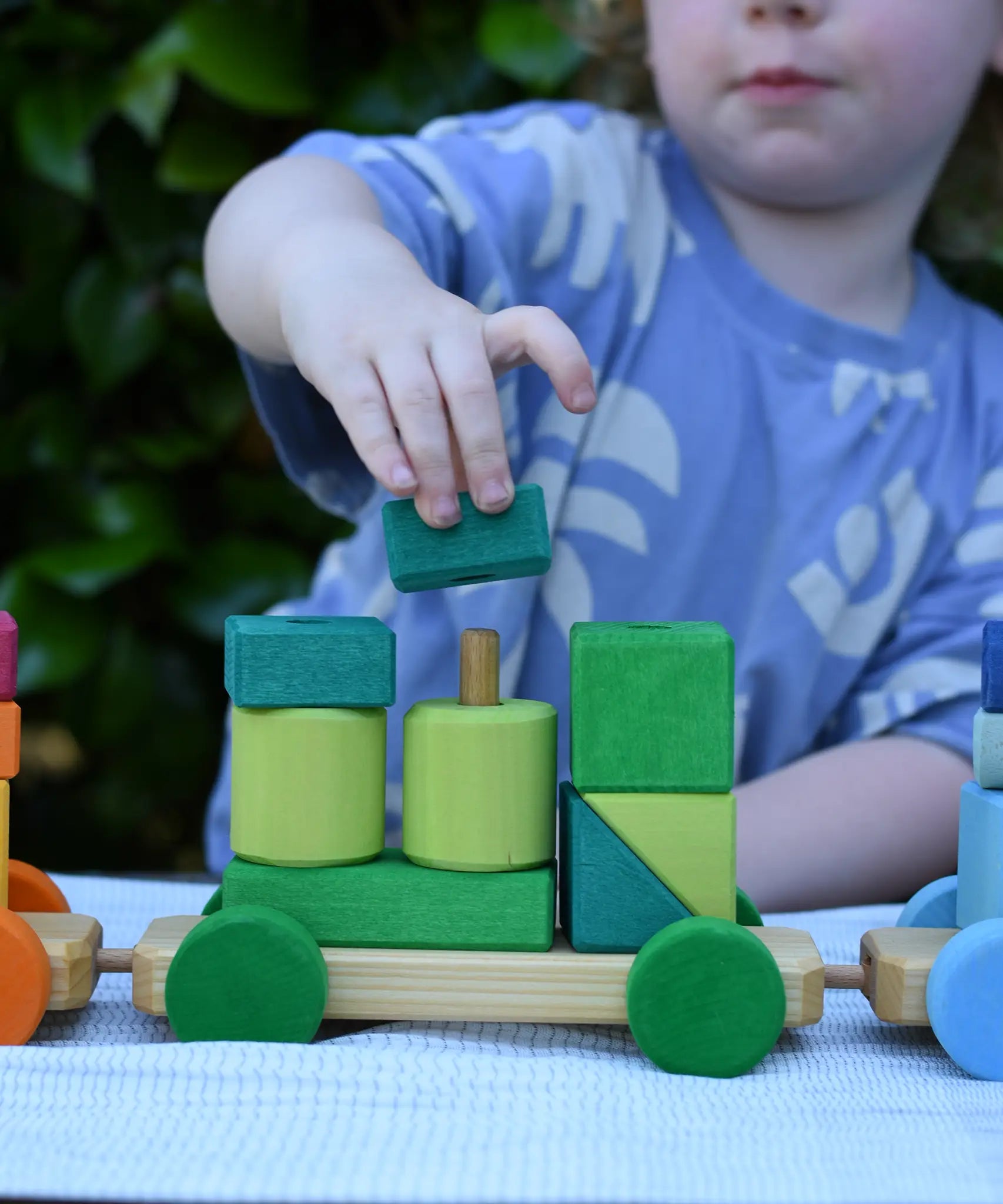 Child playing with the Glückskäfer colourful shape train wooden toy placing a square block on the pillar on the base piece.