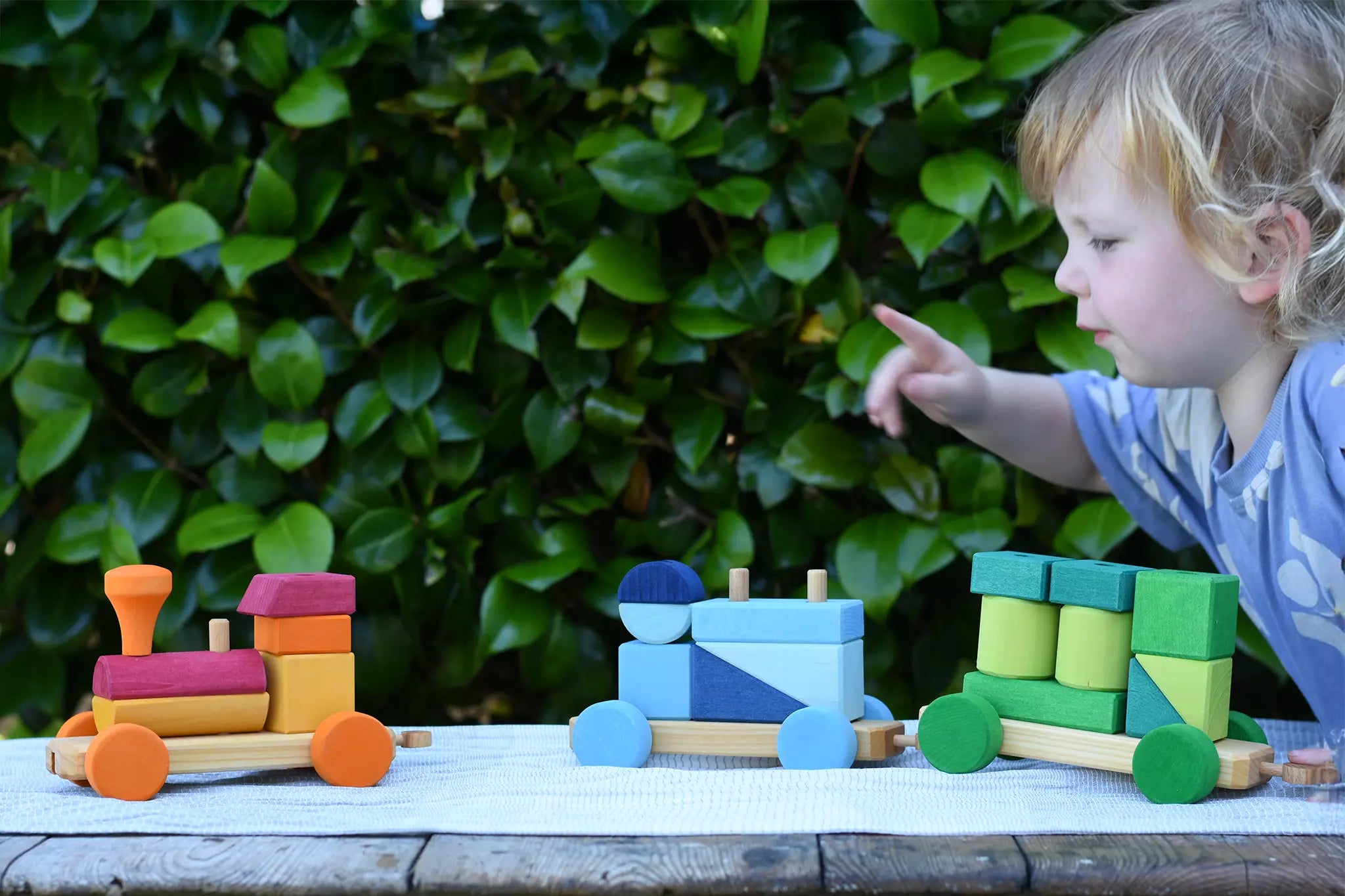 Child pointing at the Glückskäfer colourful shape train wooden toy placed on a wooden table outdoors.