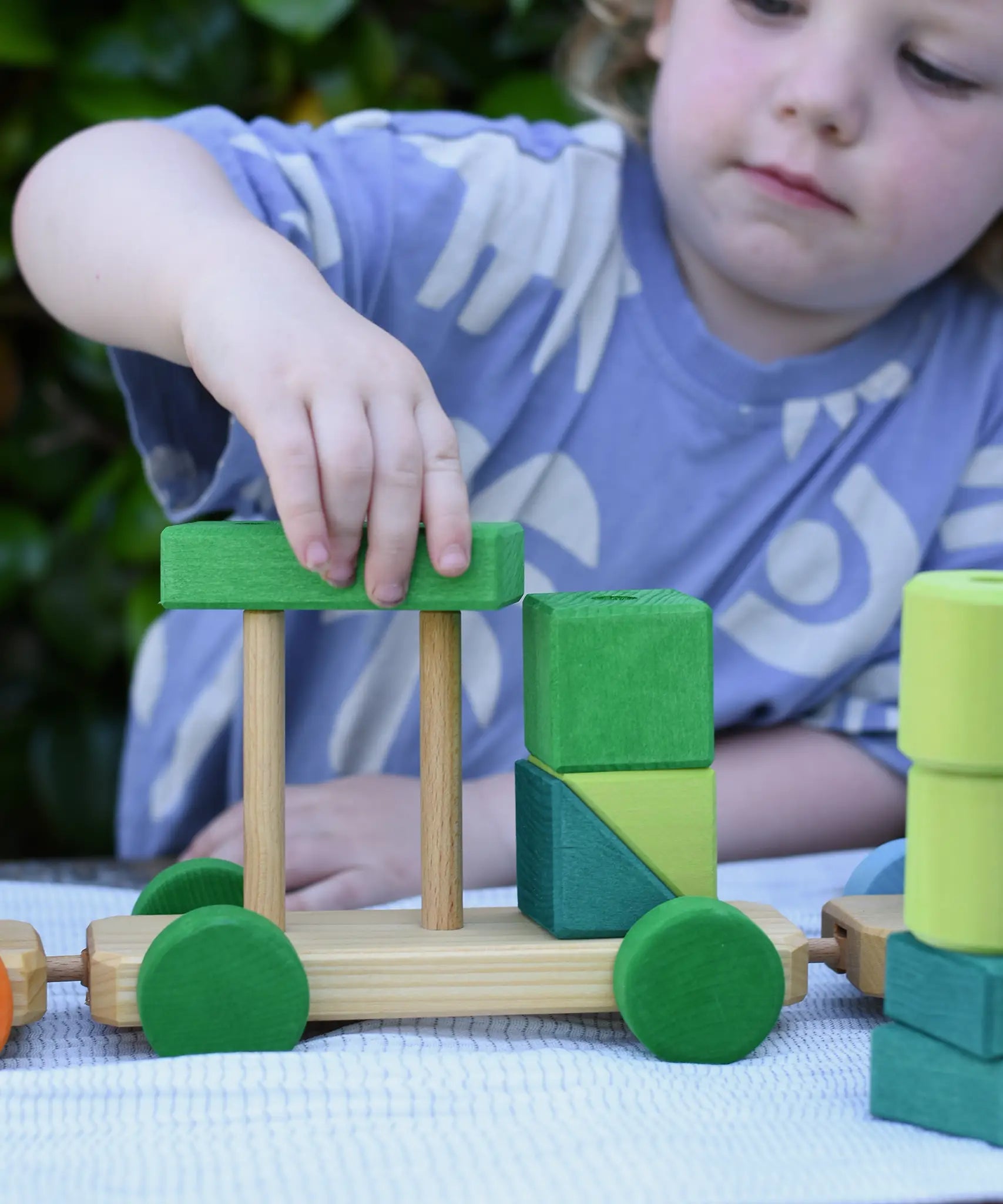 Child playing with the Glückskäfer colourful shape train wooden toy placing a green block on the pillars of the base piece.
