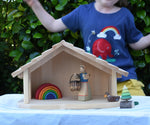 Gluckskafer farm & nativity stable wooden toy building with an Ostheimer doll, Holztiger duck and Grimm's mini rainbow placed inside. A child is dancing in the background wearing a Babipur t-shirt.