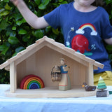 Gluckskafer farm & nativity stable wooden toy building with an Ostheimer doll, Holztiger duck and Grimm's mini rainbow placed inside. A child is dancing in the background wearing a Babipur t-shirt.