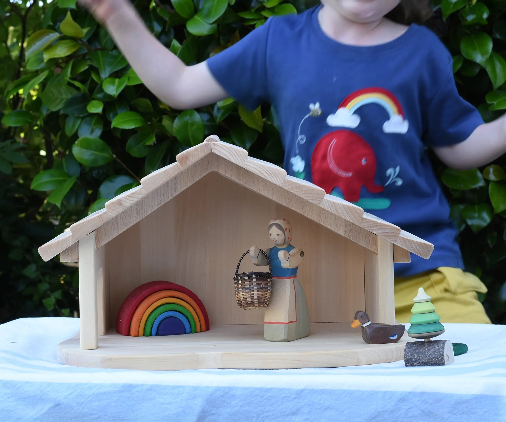 Gluckskafer farm & nativity stable wooden toy building with an Ostheimer doll, Holztiger duck and Grimm's mini rainbow placed inside. A child is dancing in the background wearing a Babipur t-shirt.