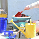 A child using a Glückskäfer Round Metal Red Shovel to prepare the soil in a pot ready to plant a Babipur seedbom in. Other Gluckskafer garden items such as a bucket and watering can can be seen at the front. 