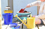 A child using a Glückskäfer Round Metal Red Shovel to prepare the soil in a pot ready to plant a Babipur seedbom in. Other Gluckskafer garden items such as a bucket and watering can can be seen at the front. 