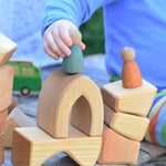 A close up of a child's hand placing a wooden Nin peg dolls on a stack of wooden blocks from the Glückskäfer 17 Large Natural Blocks Set. 