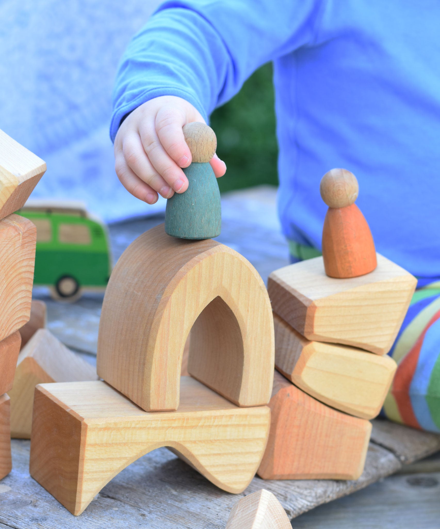 A close up of a child's hand placing a wooden Nin peg dolls on a stack of wooden blocks from the Glückskäfer 17 Large Natural Blocks Set. 