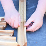 A close up of a child's hand laying out the Glückskäfer 23 Piece Wooden Marble Track laid out onto a slate surface outdoors. 