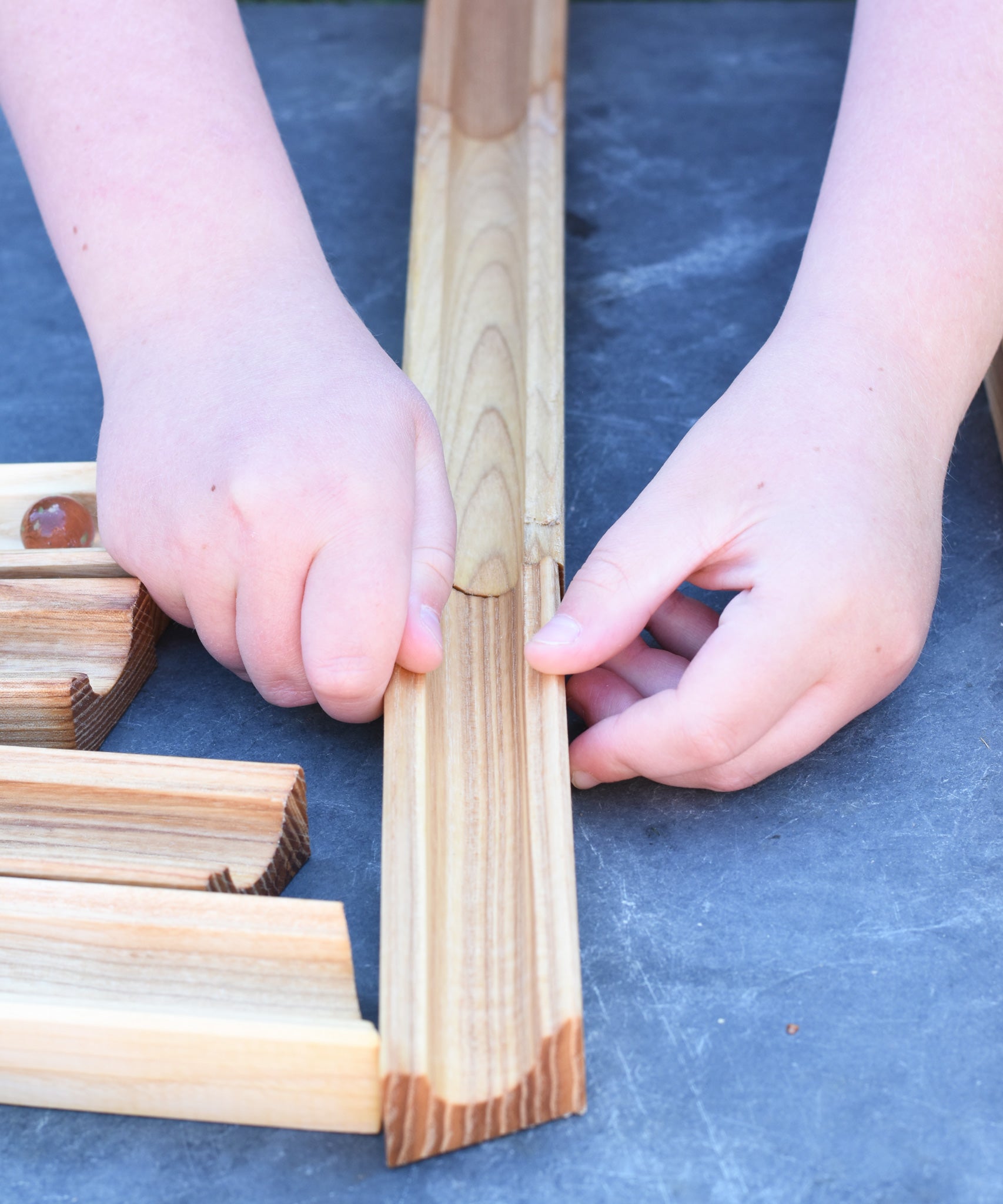 A close up of a child's hand laying out the Glückskäfer 23 Piece Wooden Marble Track laid out onto a slate surface outdoors. 