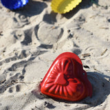 A Glückskäfer Metal red heart shaped Sand Mould placed on the sand at a beach. 