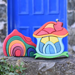 A Glückskäfer red gable house and  Mushroom House wooden stacking toy placed on a stone wall outdoors. 