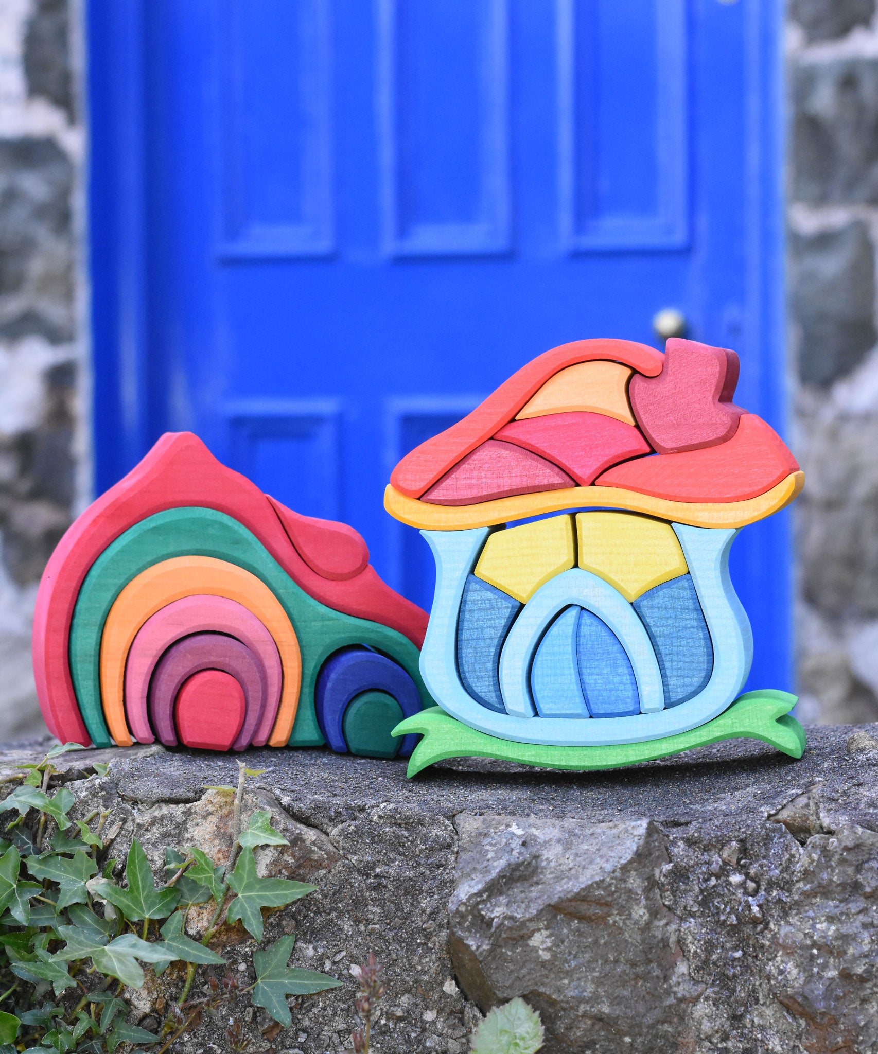 A Glückskäfer red gable house and  Mushroom House wooden stacking toy placed on a stone wall outdoors. 