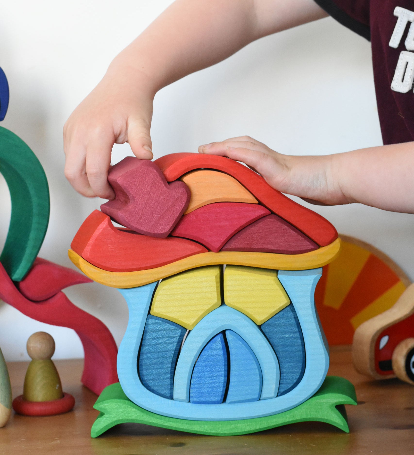 A child's hand placing the mushroom shaped chimney piece onto the Glückskäfer Mushroom House wooden toy. 