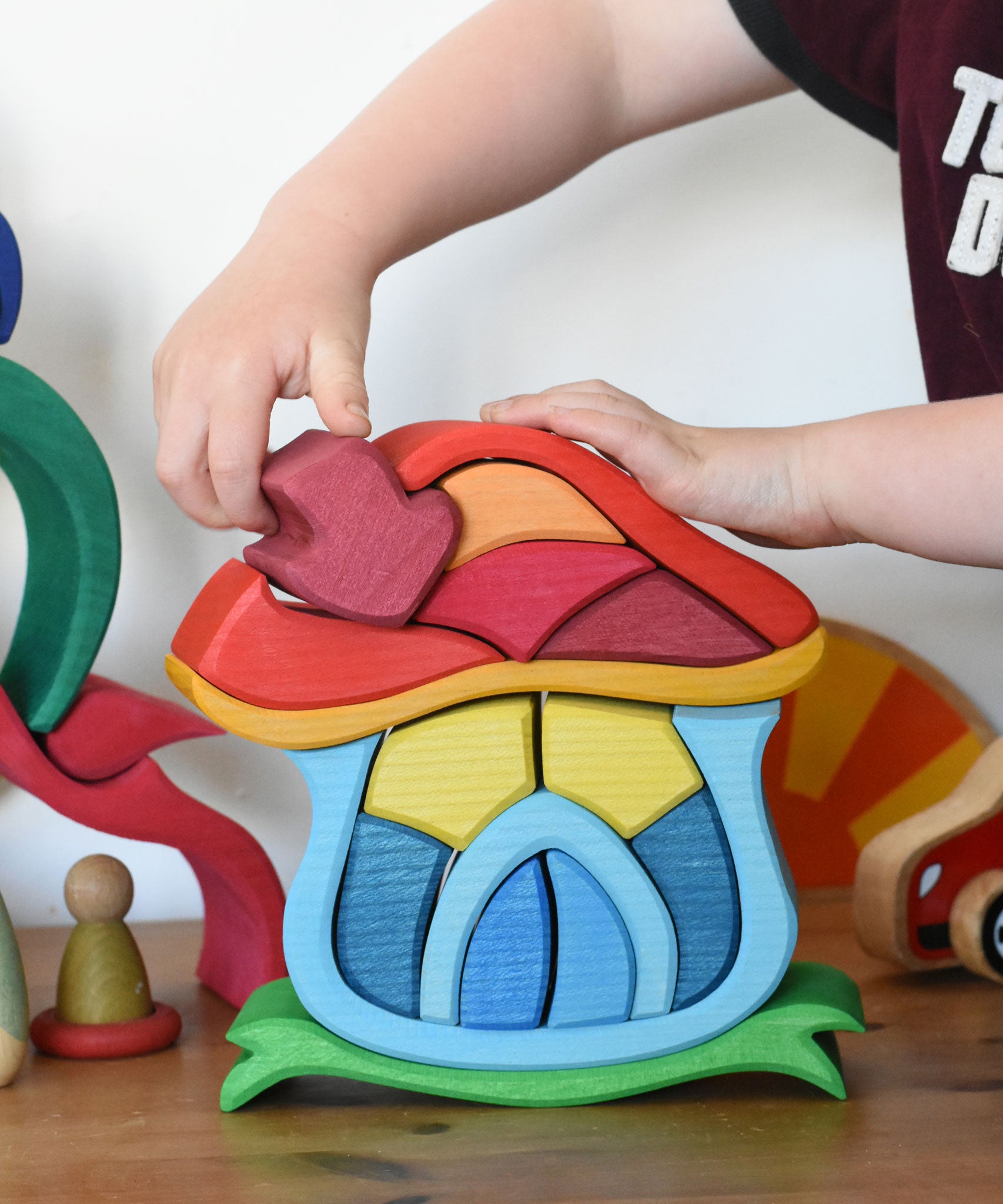 A child's hand placing the mushroom shaped chimney piece onto the Glückskäfer Mushroom House wooden toy. 
